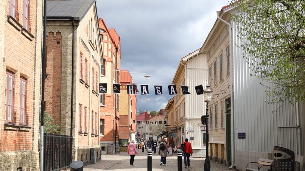Vimplar som bildar ordet Haga hänger ovan en kullerstensgata. Till vänster om gatan står tegelhus, och till höger står hus i trä.