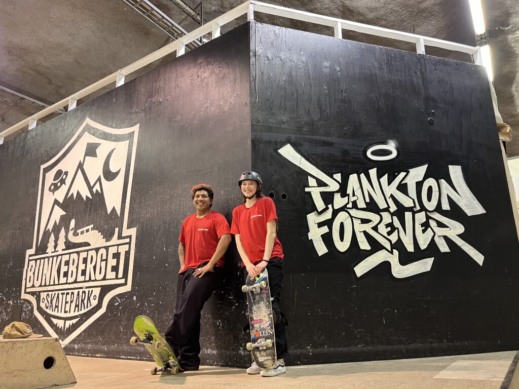 William Gonzales bredvid Vilda Tidholm, även han iklädd röd tröja. De står framför en svart vägg med texten "Bunkeberget skatepark".