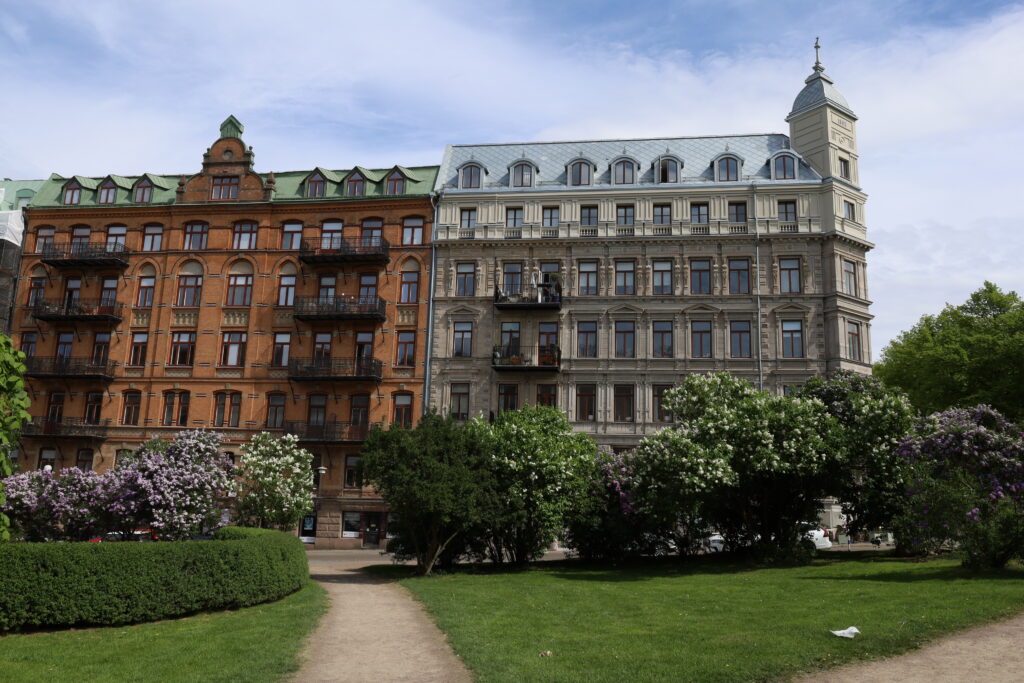 Till vänster ett hus i rödbrunt tegel och blågrönt tak, med ett fyrtiotal fönster och åtta stora balkonger. Till höger ett hus i ljust grå sten och gråblått tak, även det med ett fyrtiotal fönster. 