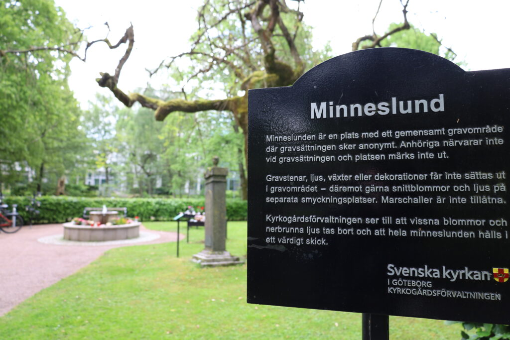 I förgrunden en svart skylt med rubriken "Minneslund", och i bakgrunden en fontän omringad av blommor.