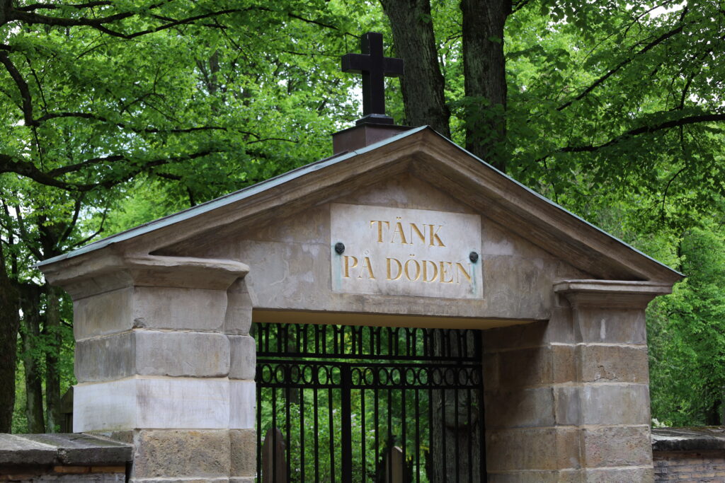 En järngrind, omringar av ett valv i sten, utgör ingången till Stampens kyrkogård. Ovanför grinden står det "Tänk på döden".