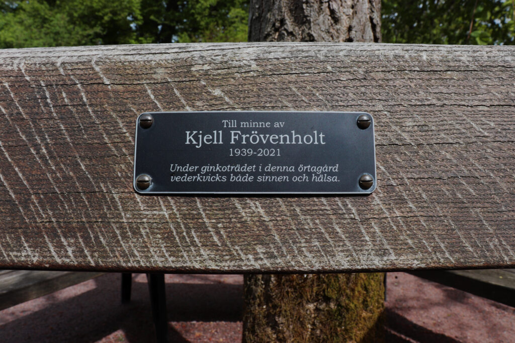 Närbild på en skylt på en bänk. Texten lyder: "Till minne av Kjell Frövenholt (1939-2021) Under ginkoträdet i denna örtagård vederkvicks både sinnen och hälsa". 