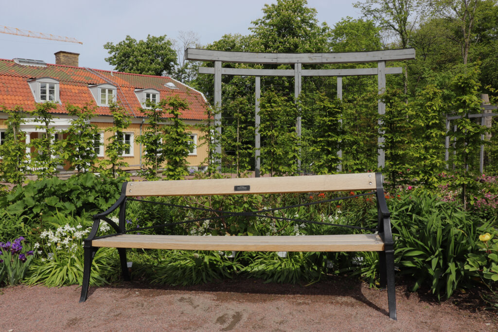 En träbänk med armstöd i svart metall, framför en rabatt med olika blomsorter. På bänkens ryggstöd sitter en plack.