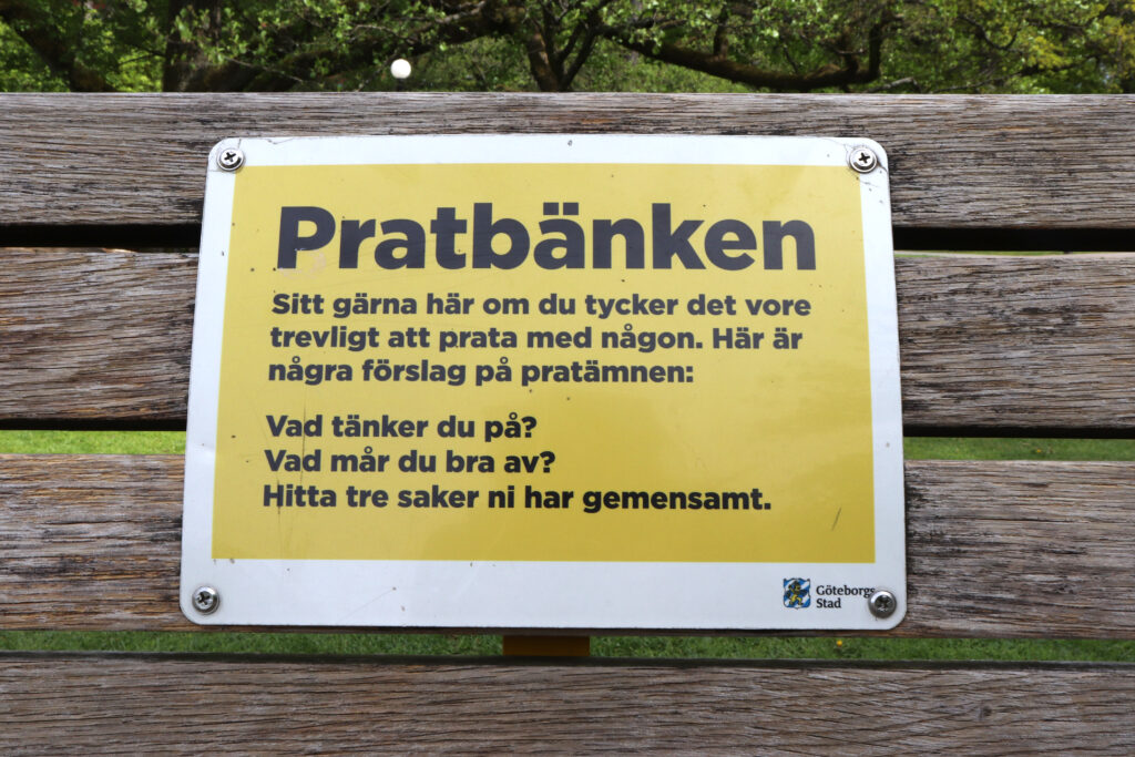 Närbild på skylten med rubriken "Pratbänk". Texten lyder: "Sitt gärna här om du tycker det vore trevlig att prata med någon. Här är några förslag på pratämnen: Vad tänker du på? Vad mår du bra av? Hitta tre saker ni har gemensamt".