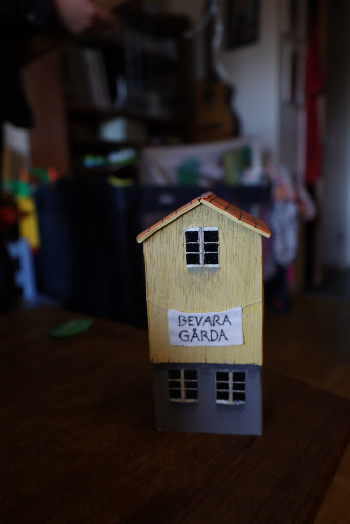 Ett gult hus med gult tegeltak och grå stengrund, med ett skynke där det står "Bevara Gårda" hängandes på fasaden. Huset är gjort av ett mjölkpaket,