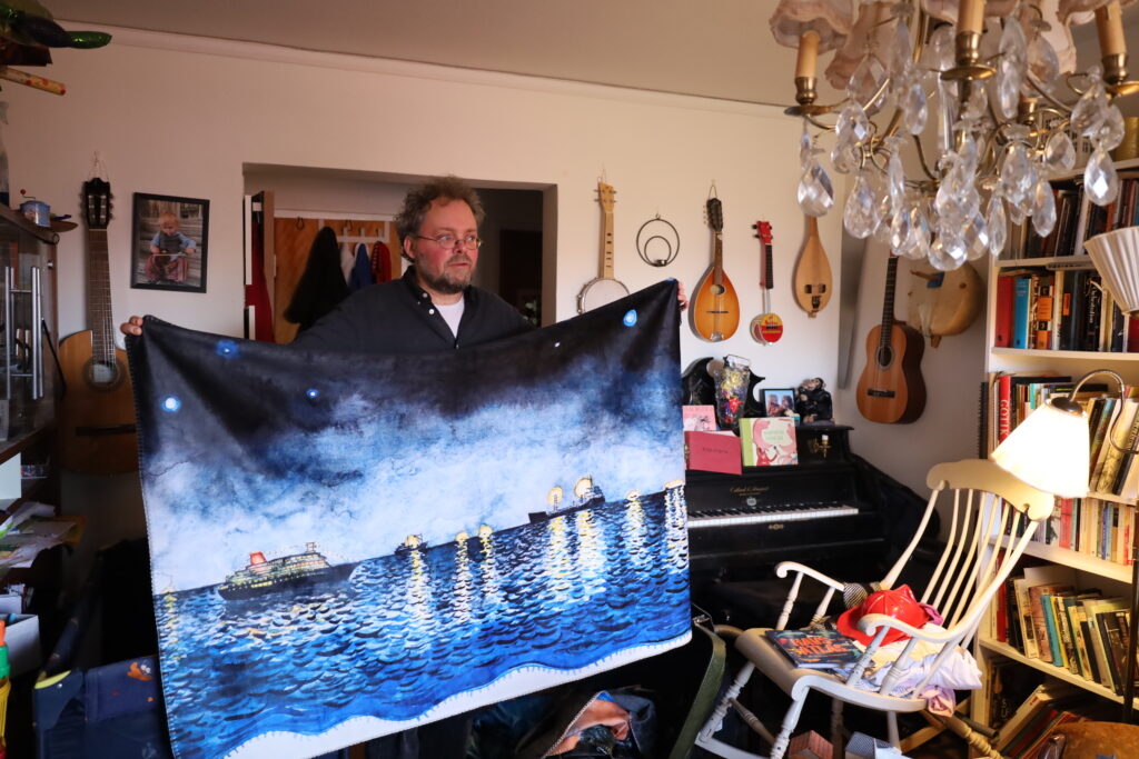 Daniel Löfberg, med en mörk skjorta och glasögon, håller upp en filt på vilken båtar på havet är målade. 