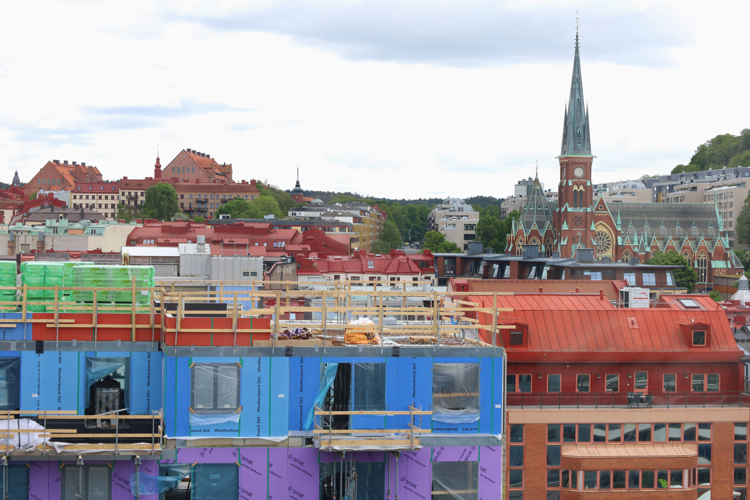 Färgglad skyddsplast på en byggnad i förgrunden. i Bakgrunden Oscar Fredriks Kyrka med sitt röda tegel och blågröna stendetaljer.
