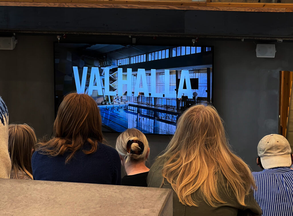 En grupp studenter sitter i en trappa och tittar på en tv-skärm. På skärmen står det Valhalla i versaler, framför en bild på hopptornet och en bassäng inne på Valhallabadet.