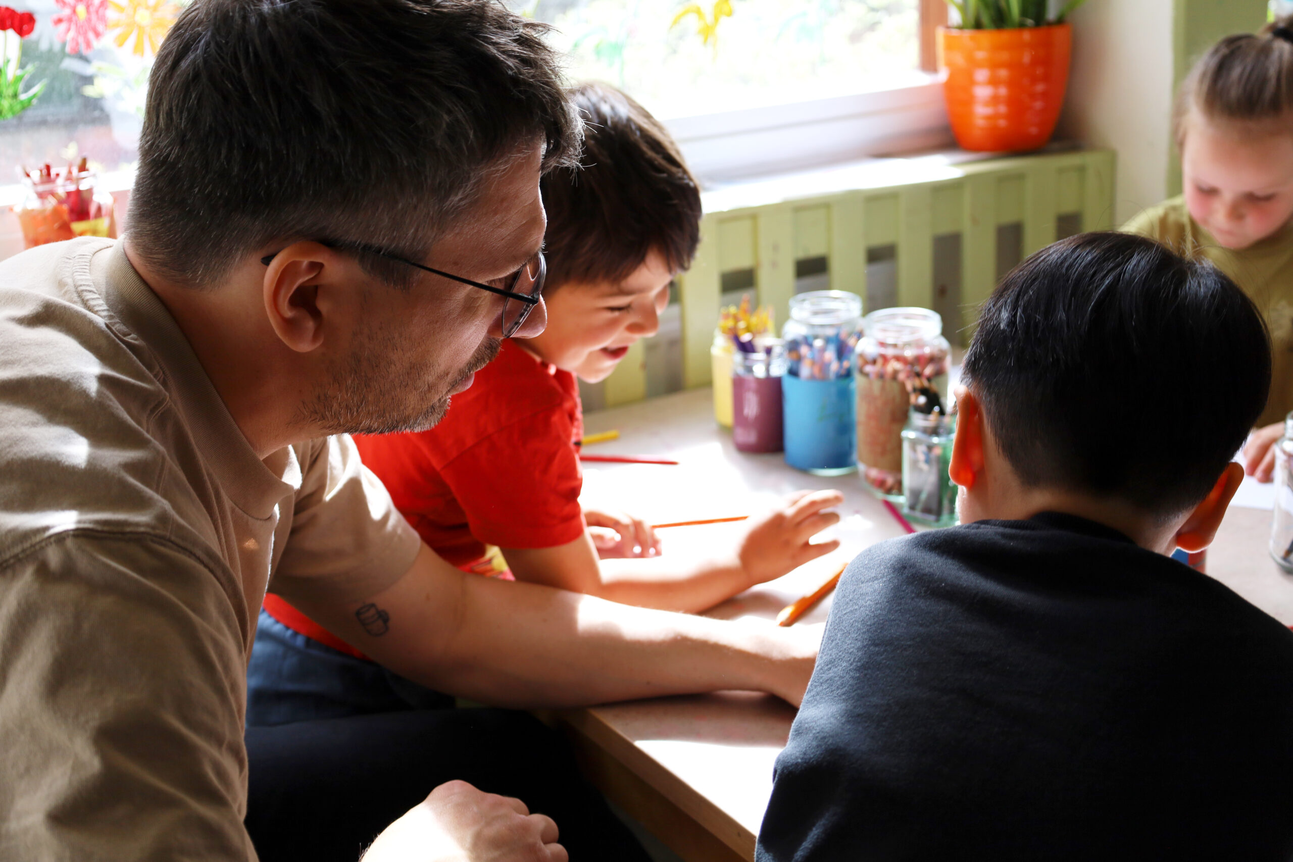 En pojke i svart tröja sitter vid bordet tillsammans med pedagogen och pojkarna i rött och grönt. De ritar tillsammans.