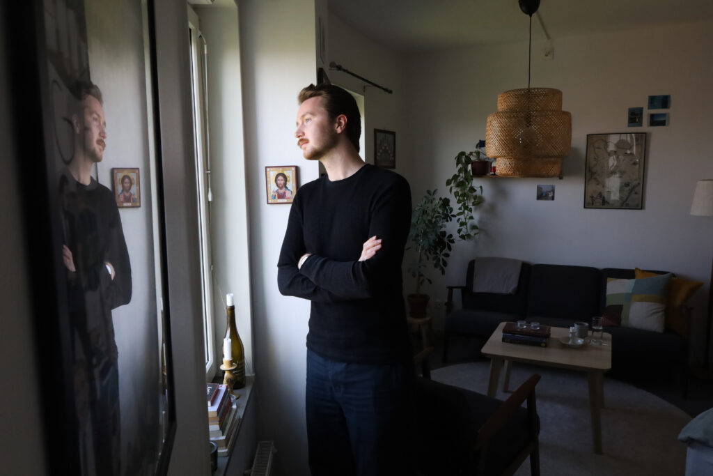 Isak Wreland står lutat mot en vägg och tittar ut genom ett fönster. Bakom honom står soffan med färgglada kuddar.