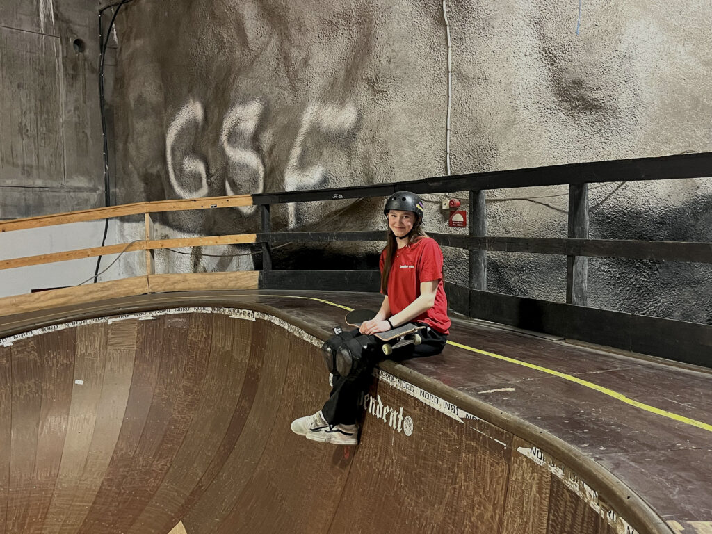 Vilda Tidholm, iklädd en röd tröja, svart hjälm och benskydd, sitter på kanten av en skateramp.  Hon har sin skateboard i knäet.