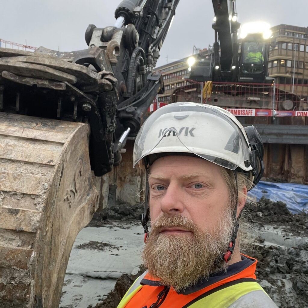 Tom Wennberg, iklädd skyddskläder, hjälm och skyddsglasögon, står framför en grävskopa.