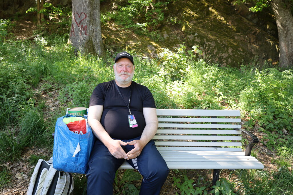 Robert Breman, iklädd svart tröja och svart keps, sitter på en vitmålad parkbänk. Till vänster om honom på bänken står en blå IKEA-kasse, och på marken bredvid bänken står en grå ryggsäck.