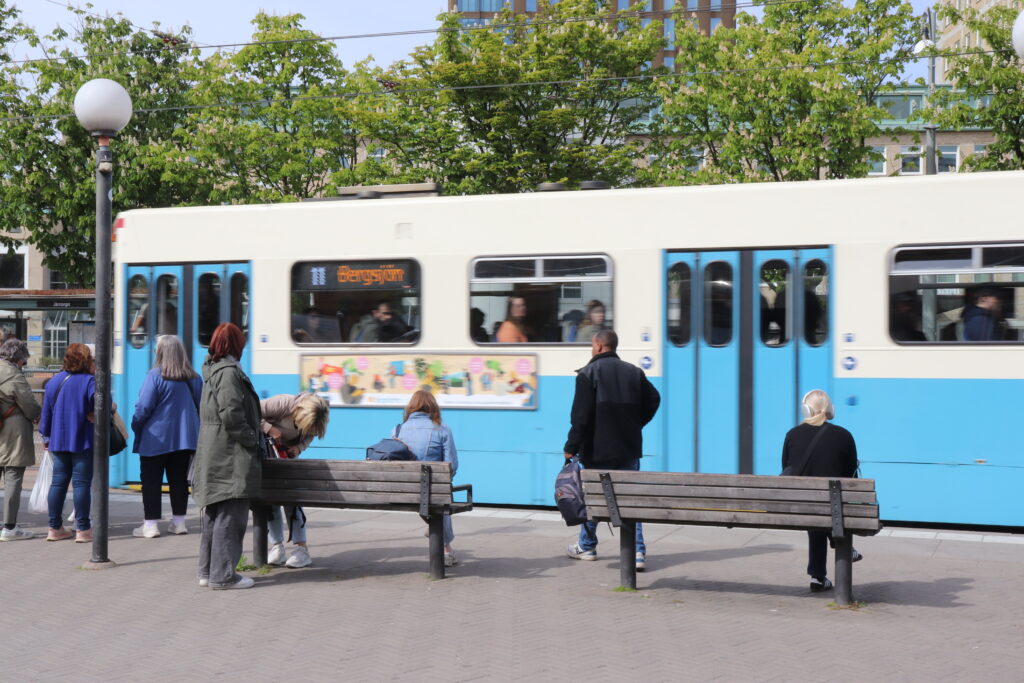 På en bänk till höger i bild sitter en blond kvinna iklädd en svart kofta. På en bänk till vänster sitter en brunhårig kvinna iklädd en jeansjacka, och bredvid henne står en blond kvinna iklädd en beige jacka och letar i en ryggsäck. Framför bänkarna åker en spårvagn förbi.